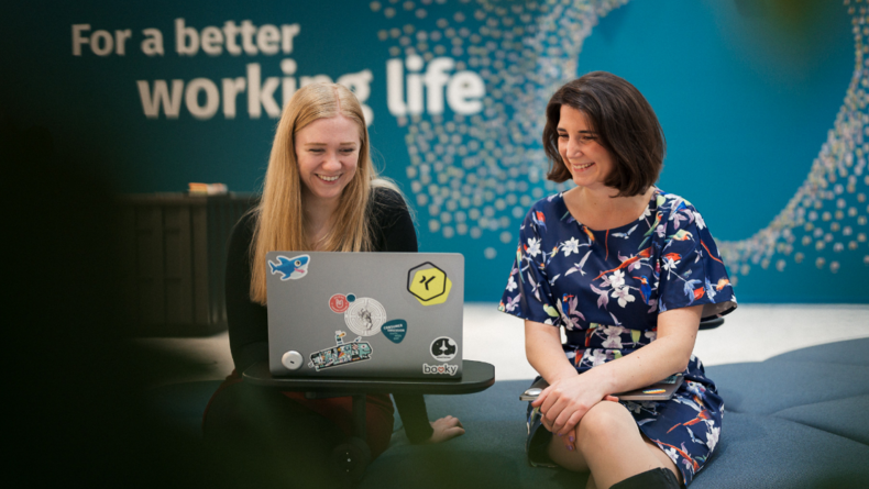 Zwei Frauen schauen gemeinsam auf einen Laptop. Im Hintergrund steht auf der Wand der Slogan "For a better working life" geschrieben.